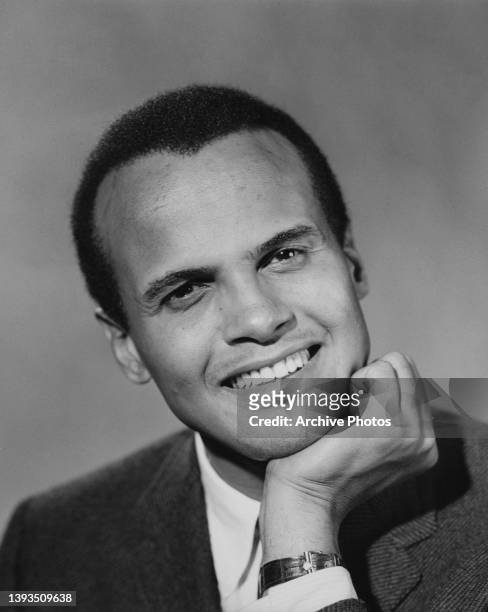 Headshot of American singer-songwriter and civil rights activist Harry Belafonte smiling with his chin resting on his left hand, an open-collar...