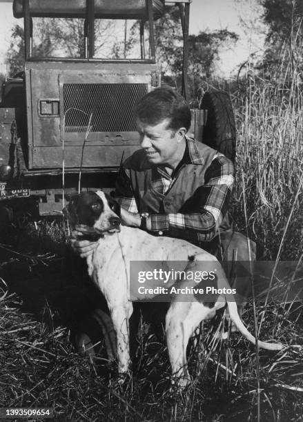 American politician Jimmy Carter crouching down to pet his dog behind an tractor on his peanut farm in Plains, Georgia, 1970. Carter served two terms...