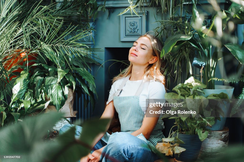 Portrait of smiling exhausted fair haired woman, seller of green shop sitting indoors near plant and flower corner. Connection with nature