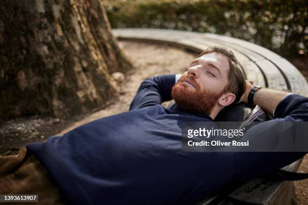 bearded young man lying on a park bench with closed eyes - tranquility stock-fotos und bilder