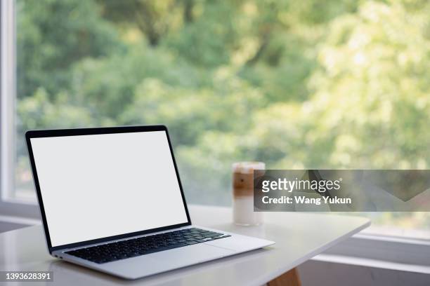 a laptop with a white screen sits on a desk stand with trees outside the window in the background - laptop on desk mockup stock pictures, royalty-free photos & images