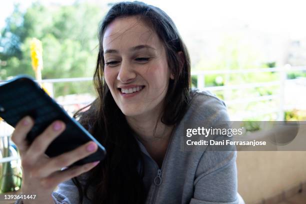young woman talking on the loudspeaker of her cell phone and smiling on the terrace of her house - flushing stock pictures, royalty-free photos & images