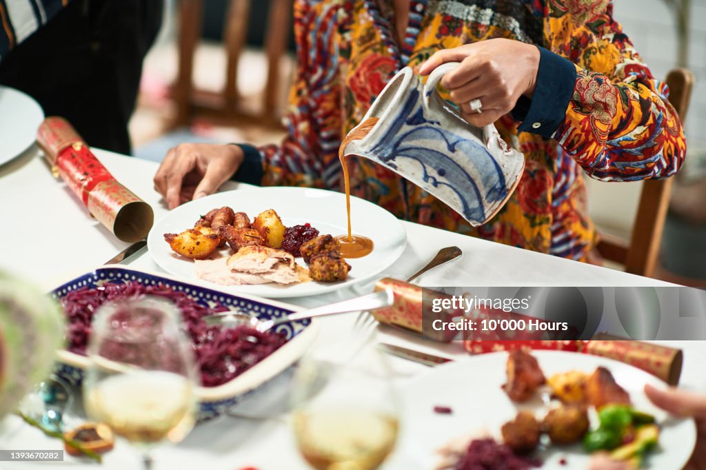 Woman pouring gravy onto roast dinner with Christmas crackers on table