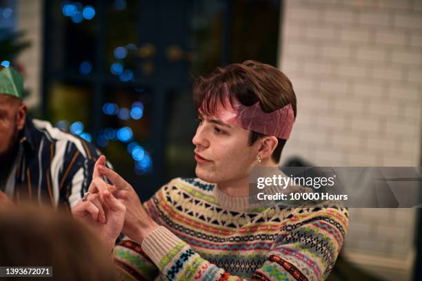 young man wearing christmas cracker hat and earring explaining and gesturing with hands - weihnachtspullover mann stock-fotos und bilder