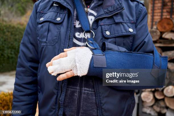 front view of man wearing bandage on injured hand while standing in front of hut, in background logs of wood ordered for use in fireplace. - personal injury fotografías e imágenes de stock