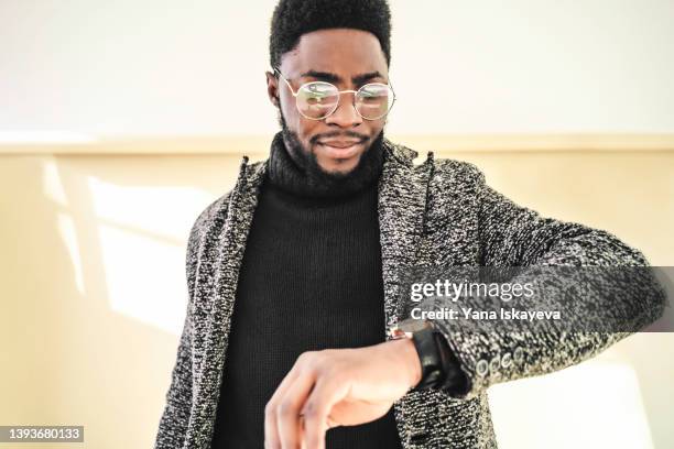 african-american hipster is looking at his wrist watches on a pastel background - mens wrist watch stock-fotos und bilder