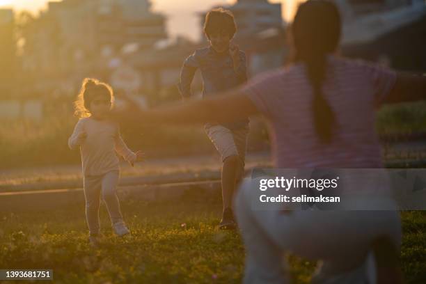 toddler daughter and elementary aged son running toward mother in public park - family with two children stock pictures, royalty-free photos & images