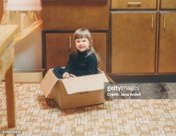 vintage child playing in cardboard box on kitchen floor, candid portrait little girl 1980s - lino stockfoto's en -beelden