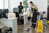 Young black man wiping computer monitors while woman with mop cleaning floor