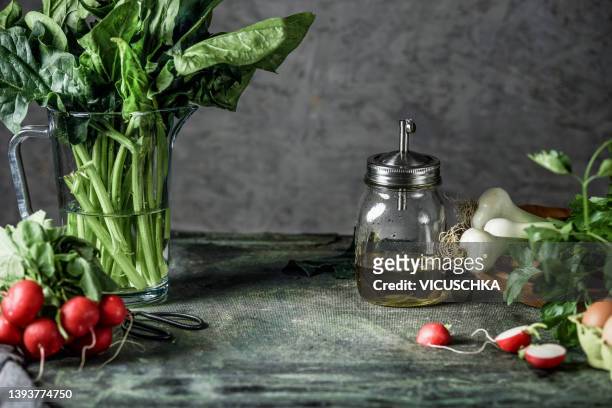 kitchen background with green spinach bunch in glass, radish, spring onion, herbs and olive oil on rustic kitchen table with dark wall background - tisch frontal stock-fotos und bilder
