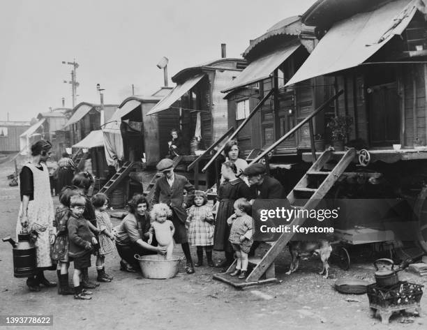 Roma woman with a large group of Roma children, one being bathed in a tin bath, with Roma caravans parked side-by-side in a row at an camp, circa...