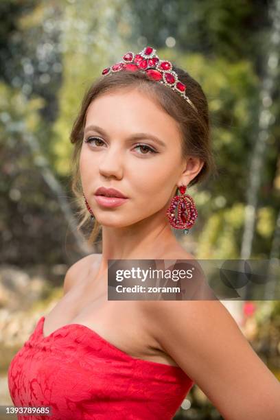 beautiful young woman in red sleeveless dress and tiara looking at camera, public park scenery - woman tiara stock pictures, royalty-free photos & images