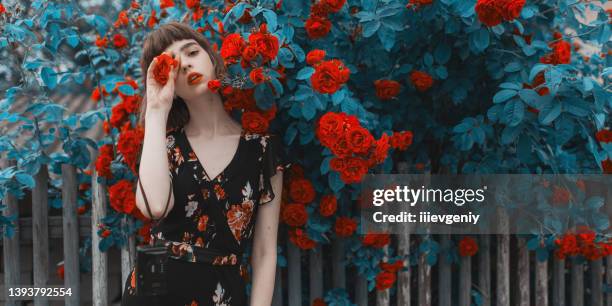 rothaariges modell auf dem hintergrund von rosenbusch. fabelhaftes asiatisches mädchen mit roten lippen im kleid auf fantastischem sommerhintergrund. rosengarten. frauenporträt. blautönung. cyan-farbe. frühlingsblumen. bob frisur - pflanzenbestandteile stock-fotos und bilder