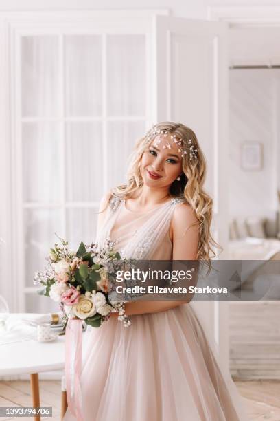 a beautiful charming young woman with hair and makeup in a long fashionable elegant dress and accessories stands and poses in a room of the indoor house. an attractive bride in a festive outfit in a wedding dress holds a spring bouquet of flowers - ärmelloses kleid stock-fotos und bilder