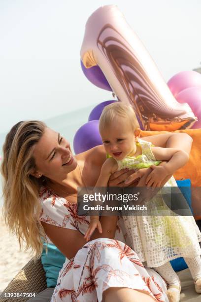 happy adorable baby girl celebrates 1 year birthday with her beautiful mother on background with decorative multicolor pillows and colorful balloons, nature background with sea beach. - number 1 mom stock pictures, royalty-free photos & images