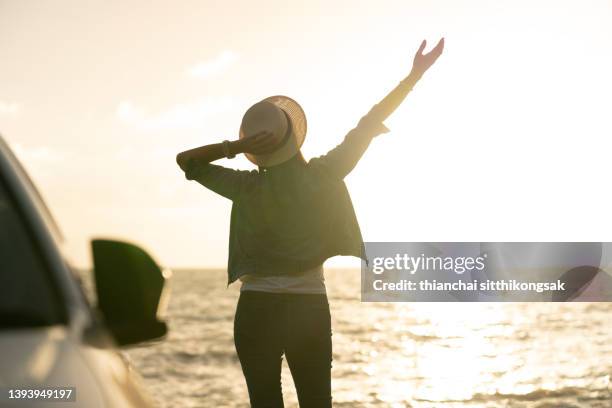 image of rear view woman on the beach with her arms outstretched beside her car in sunset - car sunset arm stock pictures, royalty-free photos & images