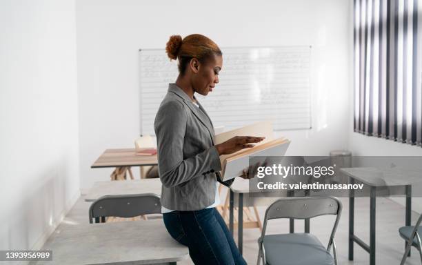 an african female teacher standing in an empty classroom and holding books - closing book stock pictures, royalty-free photos & images