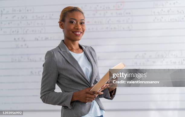 an african female teacher standing in an empty classroom and holding books - closing book stock pictures, royalty-free photos & images