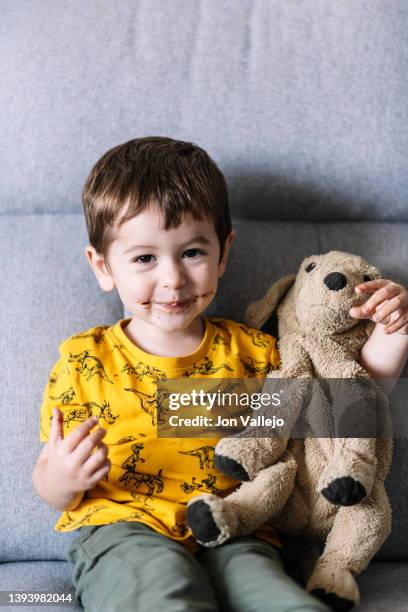 cute little boy smiling and hugging his stuffed dog while sitting on the sofa with a face full of chocolate. - leksakshund bildbanksfoton och bilder