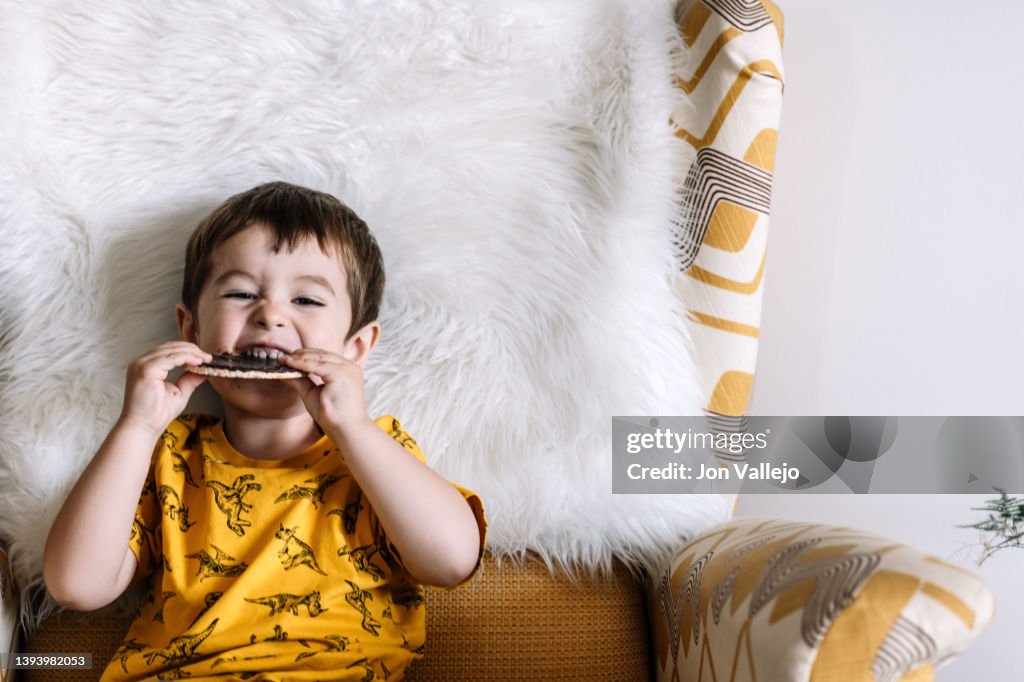 Very handsome boy sitting on a yellow sofa biting a chocolate chip cookie in his hands.