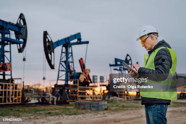 engineer in protective wear doing a project on digital tablet near nodding donkey or pump jack, oil pumps, against sunny sunset sky. - oil well foto e immagini stock