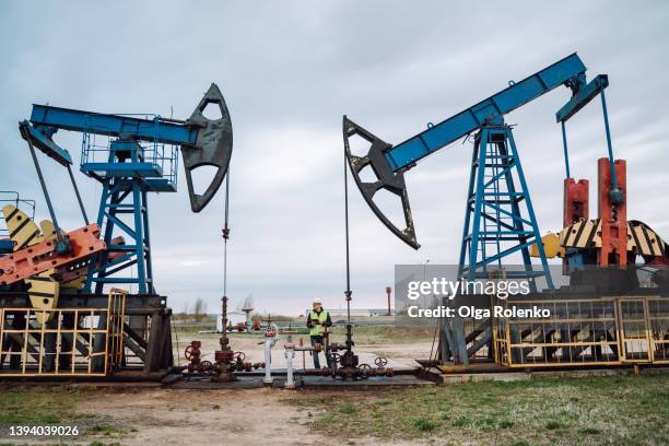 oil pumps, nodding donkey or pump jack and rig against blue cloudy sky. geologist engineer in protective wear staying in between construction - oil well stock-fotos und bilder