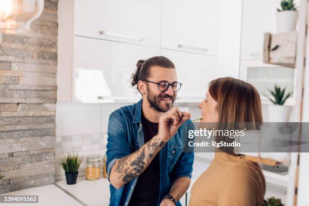 young man cooking for his girlfriend and giving her a bite of paprika - hipster in a kitchen stockfoto's en -beelden