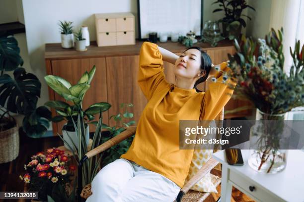 young asian woman with hands behind her head, taking a break and relaxing on armchair at cozy home, surrounded by green houseplants. lifestyle, wellbeing and wellness concept - relajacion fotografías e imágenes de stock