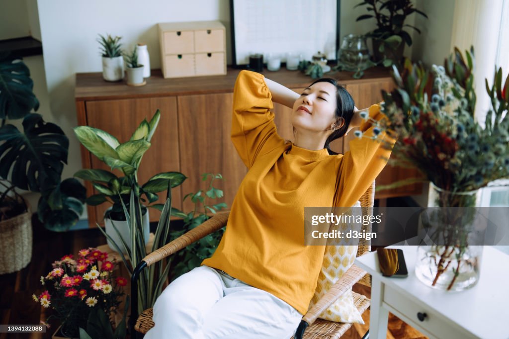 Young Asian woman with hands behind her head, taking a break and relaxing on armchair at cozy home, surrounded by green houseplants. Lifestyle, wellbeing and wellness concept