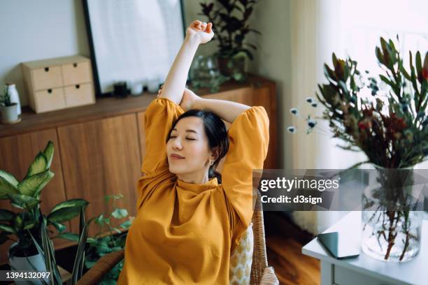 young asian woman stretching arms with her eyes closed, taking a break and relaxing on armchair at cozy home, surrounded by green houseplants. lifestyle, wellbeing and wellness concept - relief emotion stock pictures, royalty-free photos & images