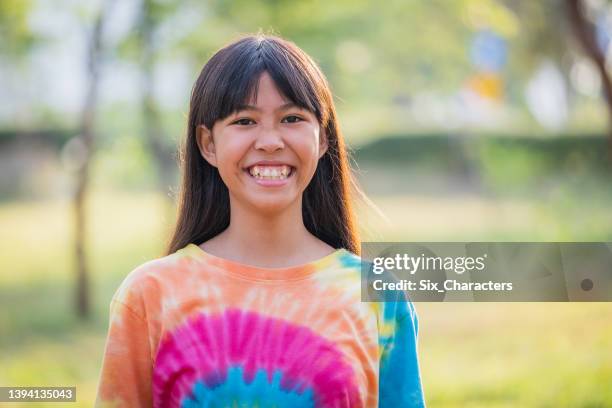 屋外の公園に立つ多色のネクタイ染料tシャツを着た若い広くスミリングアジアの女の子の肖像画 - tie dye ストックフォトと画像