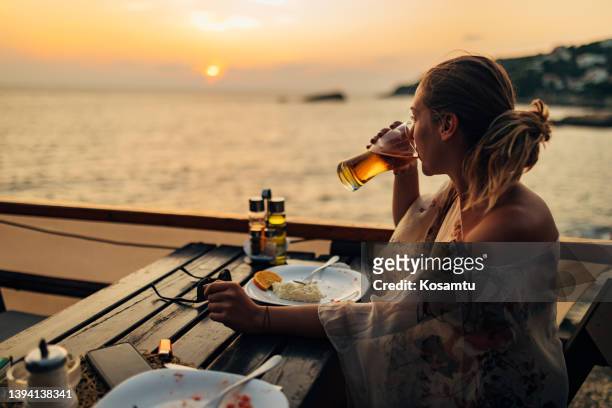 a millennial woman enjoys a golden hour while sitting in a restaurant on the beach and having lunch - eating seafood stock pictures, royalty-free photos & images
