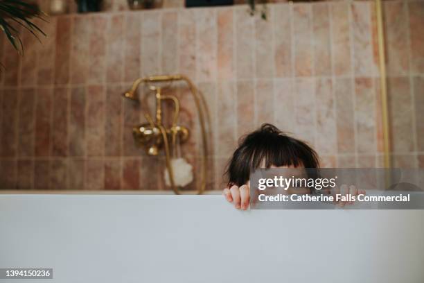 beautiful little girl peeks out of a luxurious white bath in a tiled bathroom - running water bath stock pictures, royalty-free photos & images