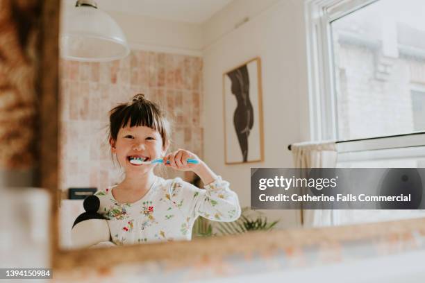 a happy little girl looks in the mirror as she brushes her teeth - lavarse los dientes fotografías e imágenes de stock