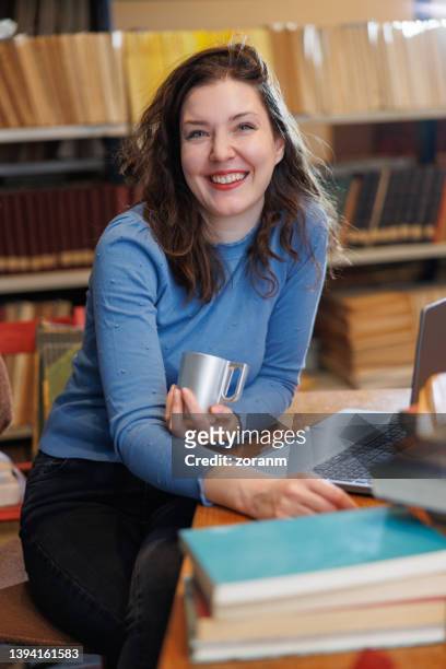 schöne frau, die am schreibtisch in der bibliothek sitzt, eine tasse kaffee hält und glücklich lächelt - librarian stock-fotos und bilder