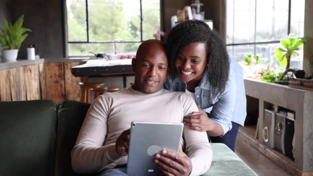 집에서 행복한 흑인 커플, 소파에서 휴식을 취하는 남자와 뒤에 서있는 여자는 말하고 웃으면서 태블릿에서 무언가를보고 있습니다.