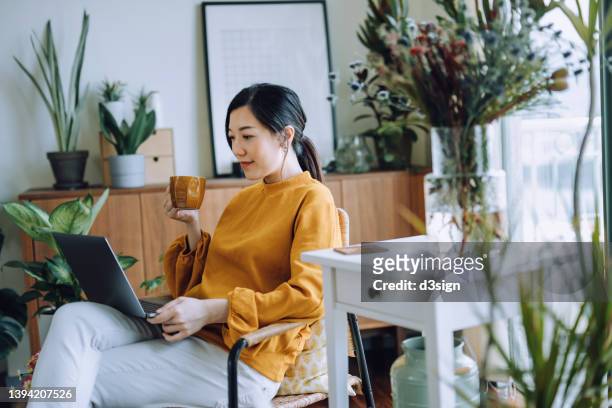 young asian woman sitting on armchair using laptop while relaxing in living room at home, surrounded by green houseplants. lifestyle and technology - conta bancária - fotografias e filmes do acervo