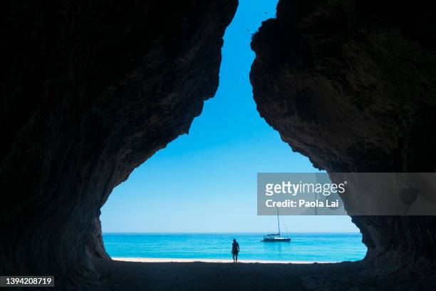 a sailboat by the coastline in a turquoise sea, seen from a cave, with a person walking on the beach. - sardinien stock-fotos und bilder