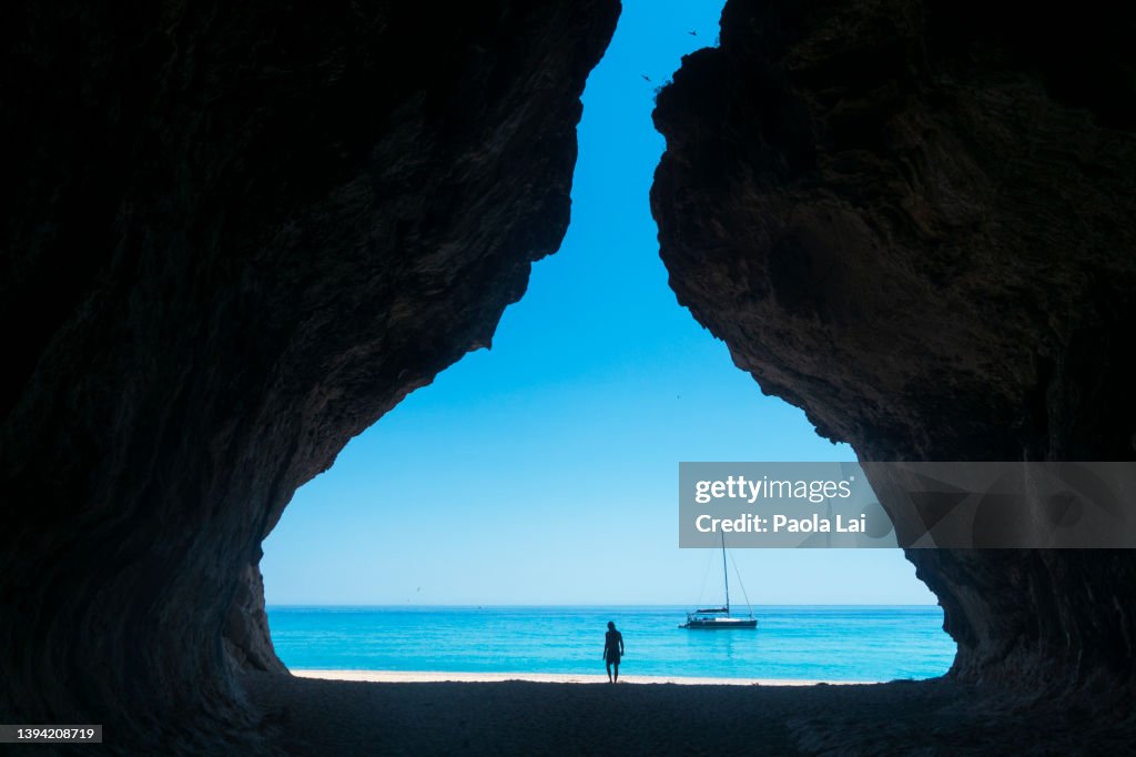 A sailboat by the coastline in a turquoise sea, seen from a cave, with a person walking on the beach.