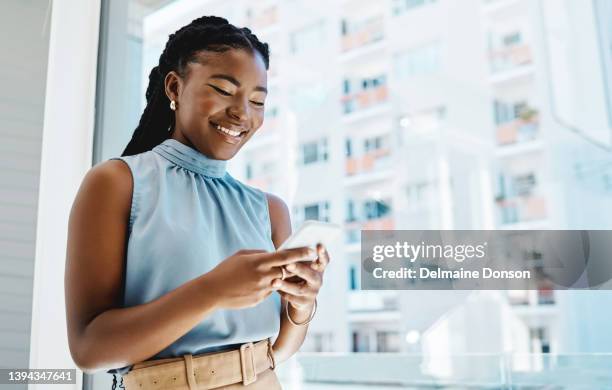 happy young black businesswoman using a cellphone while standing at a window in an office alone - africa phone black stock pictures, royalty-free photos & images