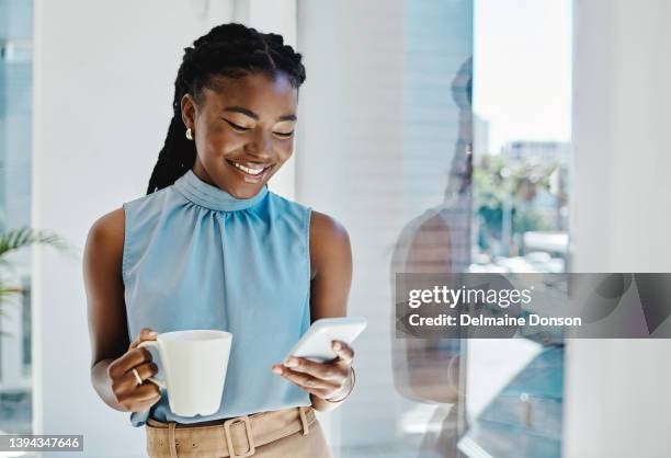 happy african businesswoman drinking coffee while texting on a cellphone at a window in an office alone - black mug stock pictures, royalty-free photos & images