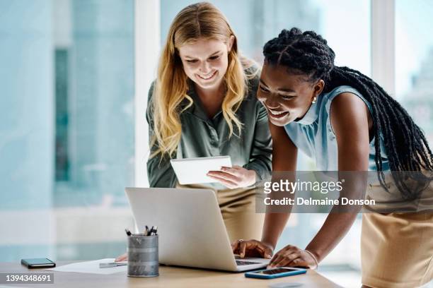 dos mujeres de negocios diversas que trabajan juntas en una tableta digital y una computadora portátil en una oficina - jovenes felices fotografías e imágenes de stock