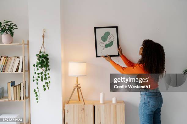 young woman with curly hair hanging picture frame on wall at home - finishing touch stock-fotos und bilder