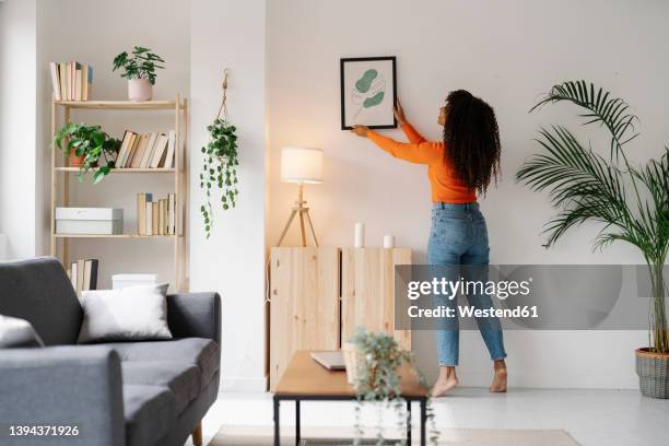 young woman hanging picture frame on wall in living room at home - hanging photos stock pictures, royalty-free photos & images