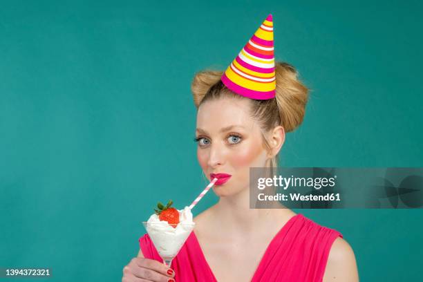 young woman in party hat drinking strawberry cocktail with straw against green background - adult cocktail party background stock-fotos und bilder