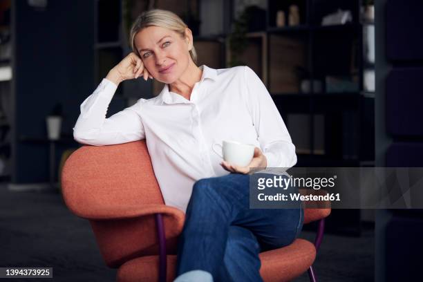 smiling businesswoman holding coffee cup sitting with head in hand on chair - überkreuzte beine stock-fotos und bilder