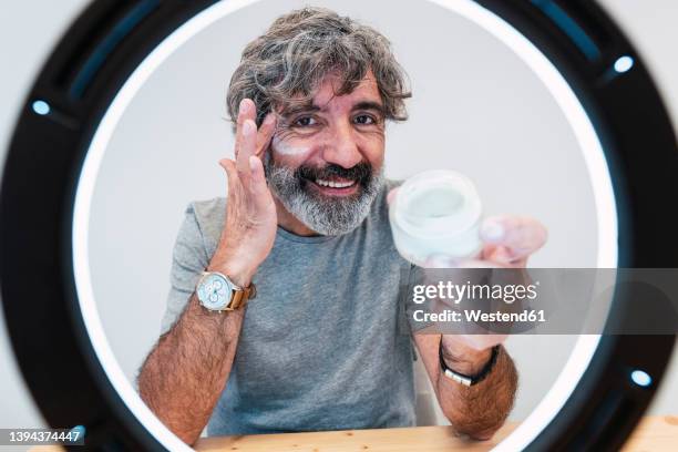 smiling senior man applying moisturizer on face seen through ring light at home - beauty influencer stock pictures, royalty-free photos & images