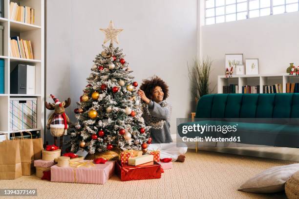 smiling young woman sitting cross-legged decorating christmas tree at home in living room - house presents golf day inside foto e immagini stock