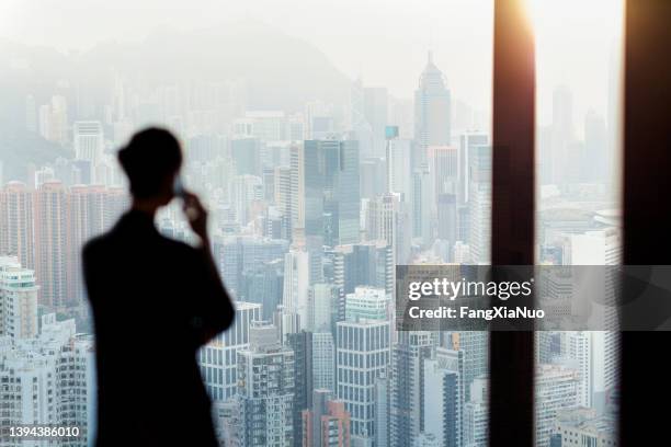 businesswoman talking on phone from office window overlooking hong kong cityscape - clinton global initiative addresses issues of worldwide concern stockfoto's en -beelden