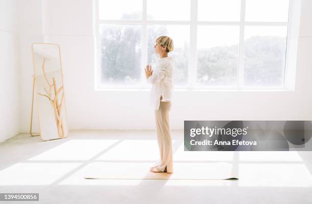 beautiful authentic woman with a short blond hair is preparing for a yoga practice on a yoga mat in front of a window. she is wearing a light-colored casual clothing. concept of relaxation exercises - good posture 個照片及圖片檔